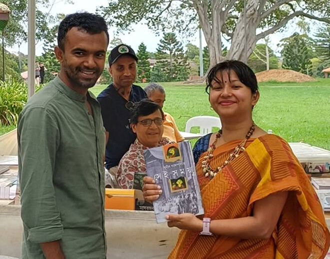 Anandodhara members at their book stall holding a Bengali publication, outdoors