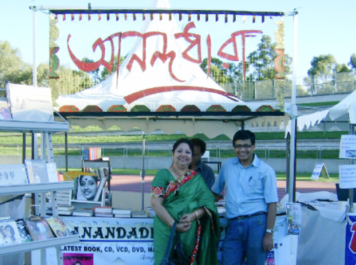 Anandodhara outdoor book stall under a tent, with two members in front of the display