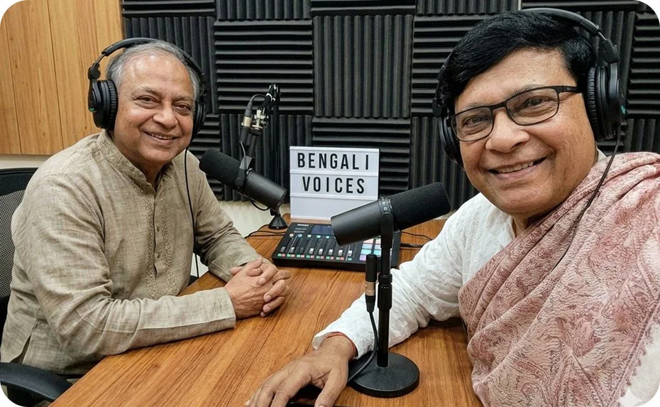 Two Anandodhara podcast hosts wearing headphones at a studio desk with microphones and a lightbox sign reading Bengali Voices